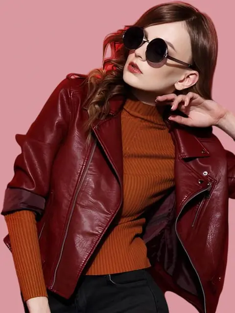 Woman wearing a burgundy leather jacket over an orange knit turtleneck, with round sunglasses against a pink backdrop, posing in a fashion shot.