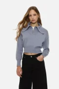 Young woman wearing a light blue cropped knit sweater with a large curved collar and front zipper, paired with black jeans against a white background.