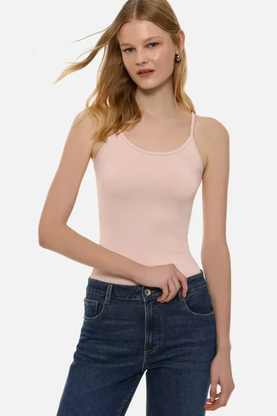 Woman wearing a pale pink sleeveless camisole and dark blue jeans, posing with one hand at her waistband against a white background.