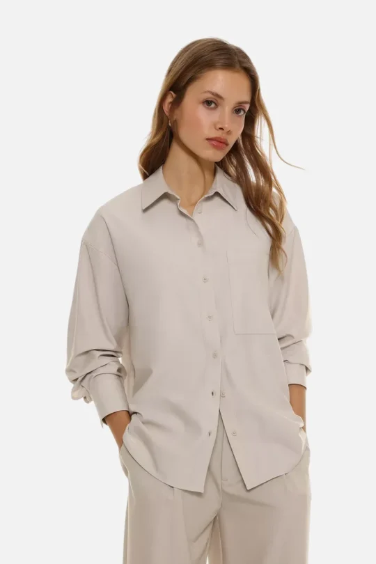 Model wearing an oversized beige button-up shirt with a chest pocket and matching wide-leg pants, hands in pockets against a white background.